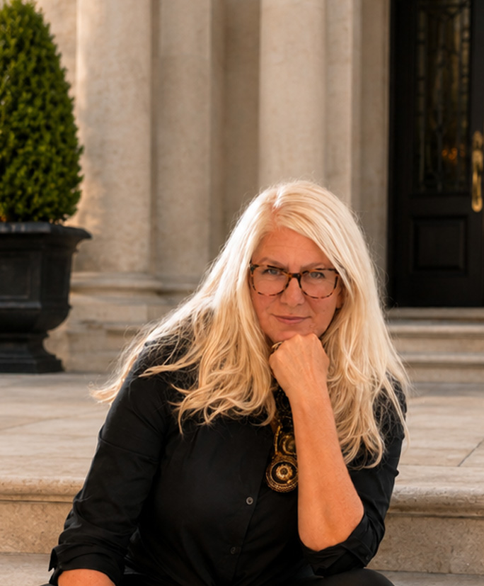 Sandi Wilen seated on the stone steps of a grand home entrance with columns and topiaries.